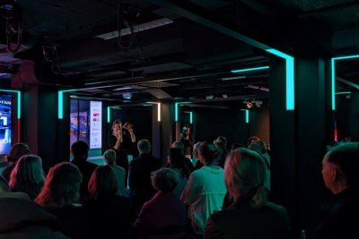 Viele Personen sitzen zur Bühne gewandt, auf der eine Frau spricht