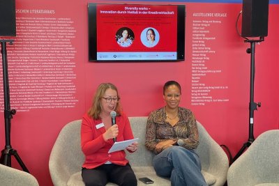 Zwei Frauen sitzen auf einer Bühne. Die linke Frau mit Mikro.