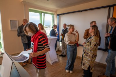 Eine Gruppe von Personen hält sich in einem Raum auf, einzelne Personen unterhalten sich, eine Frau im Vordergrund des Bildes guckt sich Fotos an