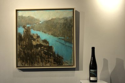 An einer weißen Wand hängt ein Gemälde einer Flusslandschaft. Daneben steht eine Weinflasche auf einem Ständer