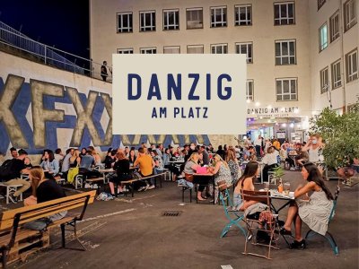 In einem Hinterhof sitzen viele Leute an Tischen und unterhalten sich. In der Mitte des Bildes sieht man die Überschrift &quot;Danzig am Platz&quot;
