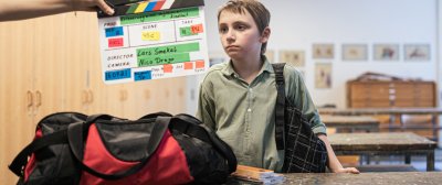 Ein Kind steht in einem Klassenzimmer vor einem Tisch mit einer Sporttasche. Eine Filmklappe wird vor ihn gehalten