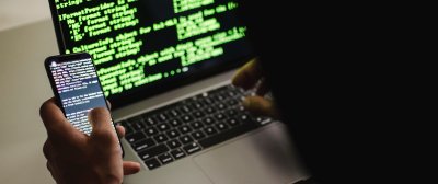 Eine Person sitzt am Laptop und hält ein Smartphone in der Hand. Auf dem Laptop ist ein Programmierskript zu sehen