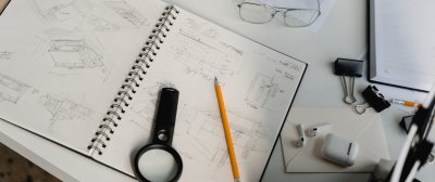Man sieht den Tisch eines Architekten, auf dem Tisch sind Skizzen von Gebäuden, Bleistifte, eine Lupe etc. zu sehen